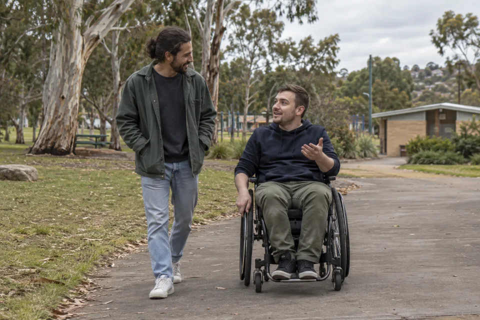 A wheelchair user and a companion talk on a suburban park path.